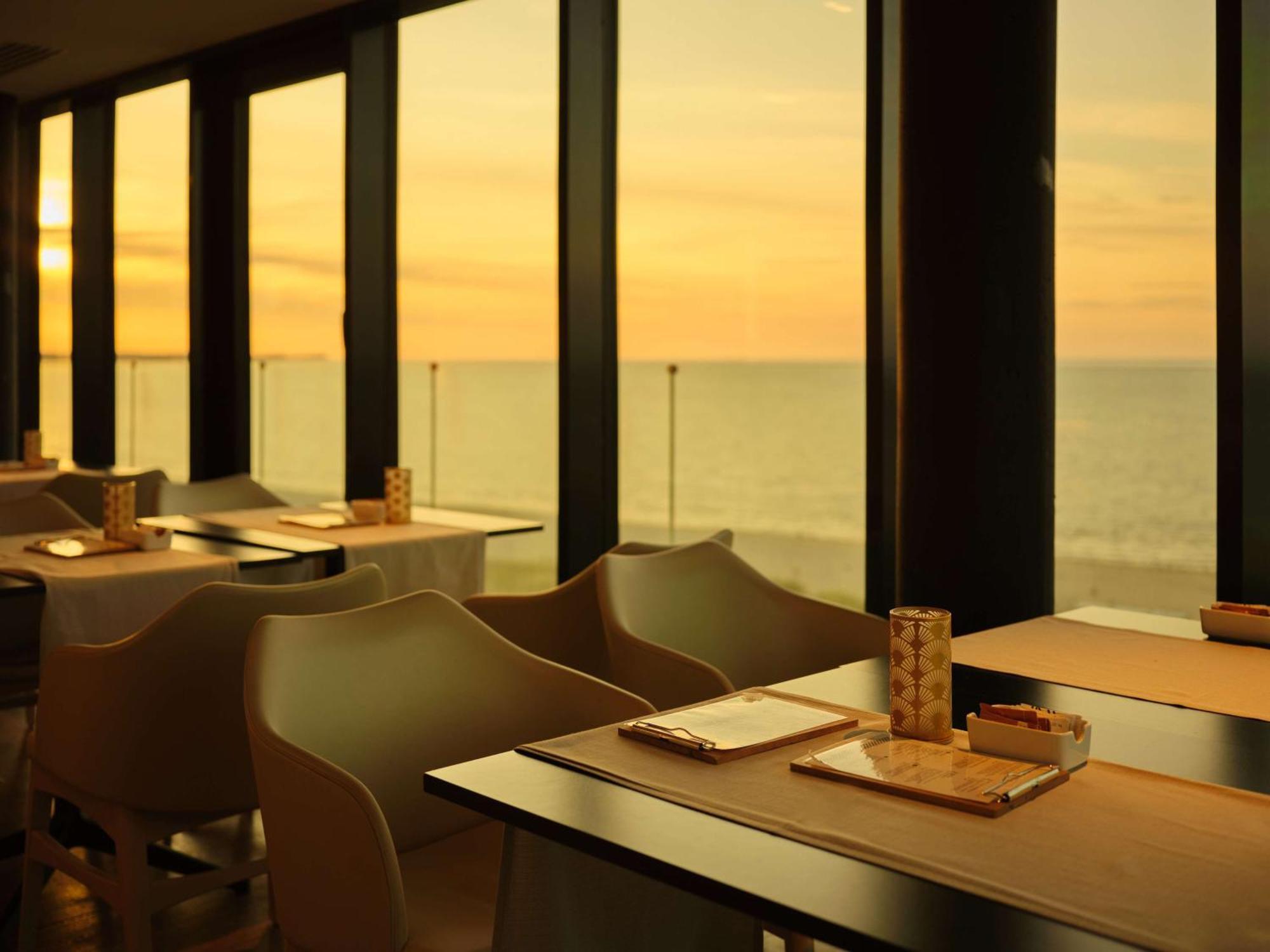 Radisson Blu Resort Swinoujscie Ngoại thất bức ảnh The photo shows a restaurant interior with a beautiful view of the ocean during sunset. The large windows allow natural light to flood the space, highlighting the warm colors of the sunset in the sky. There are several tables, each set with placemats