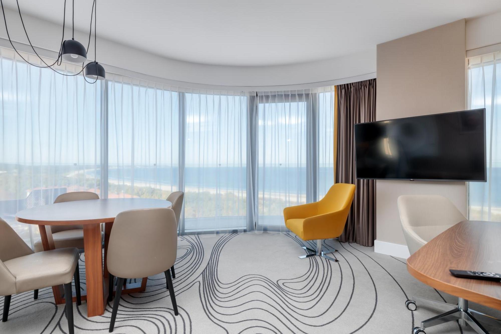 Radisson Blu Resort Swinoujscie Ngoại thất bức ảnh The image shows a modern hotel room or suite with a bright, airy atmosphere. There are large windows that allow natural light to fill the space, providing a view of the ocean. The decor includes a round dining table with several chairs, and a stylish