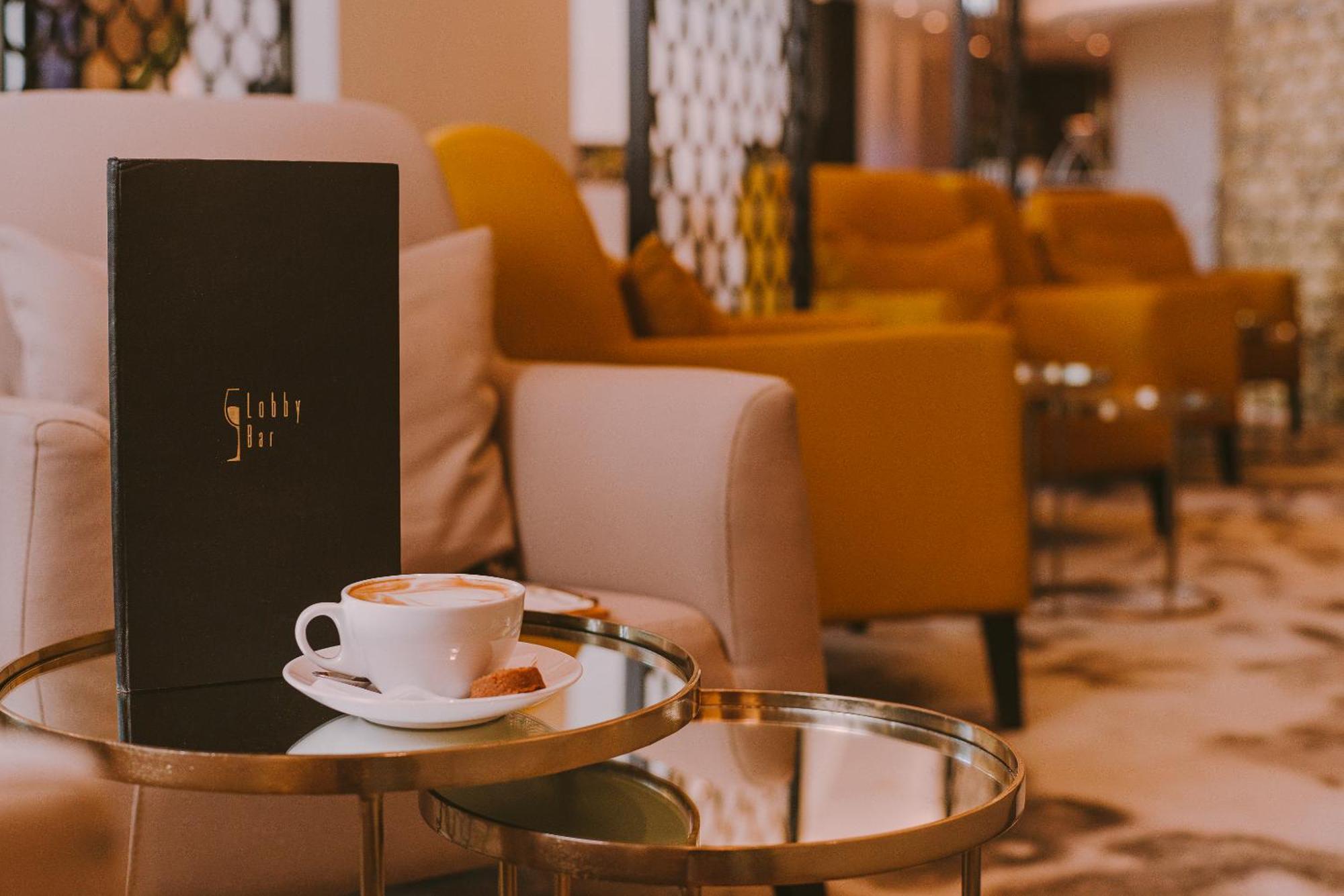 Radisson Blu Resort Swinoujscie Ngoại thất bức ảnh The photo shows a cozy indoor setting with a stylish, modern atmosphere. In the foreground, there is a small round glass table with a white coffee cup and a saucer, alongside a small biscuit or cookie. Next to the coffee, there is a black menu with e
