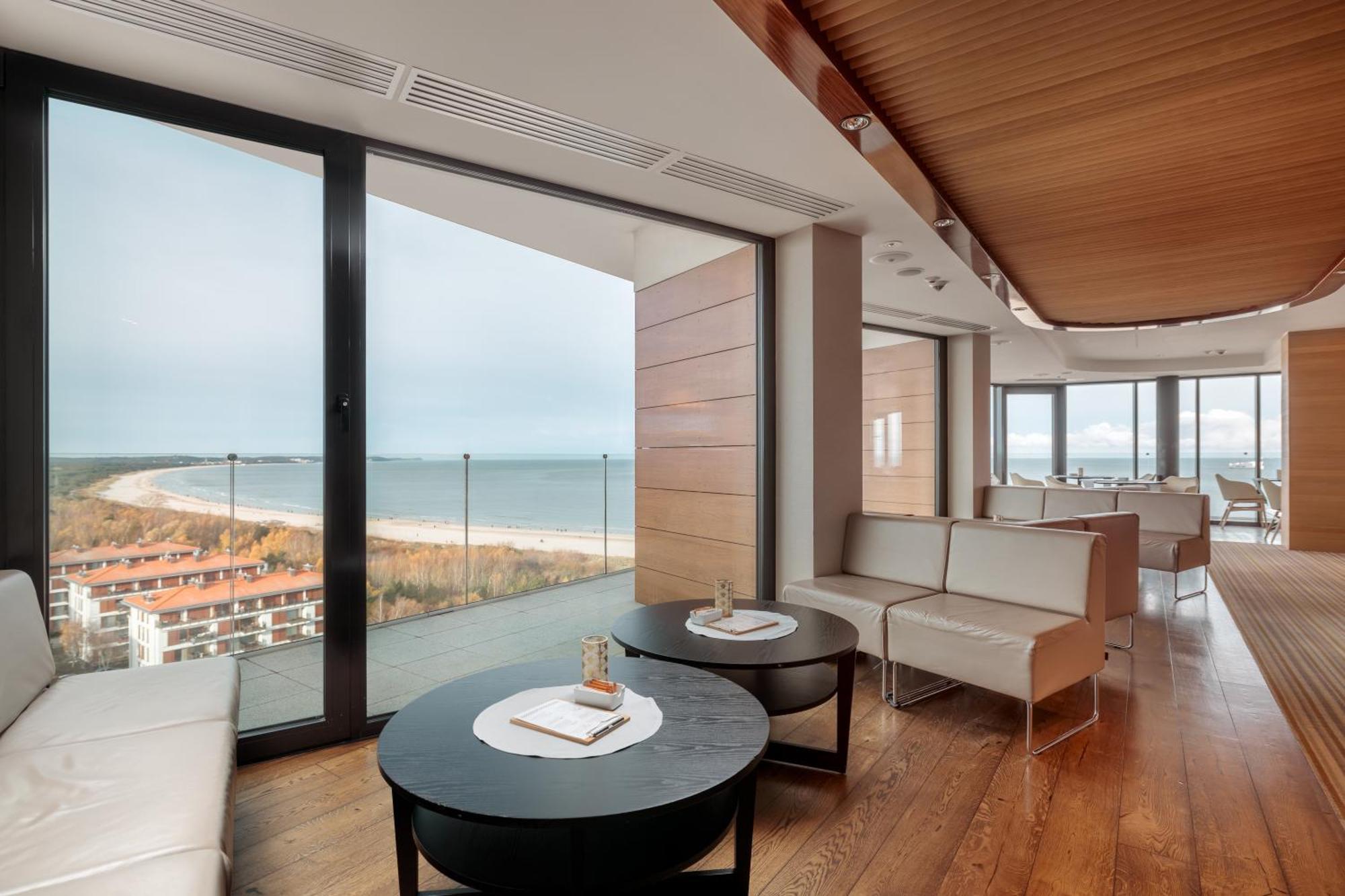 Radisson Blu Resort Swinoujscie Ngoại thất bức ảnh The photo shows a modern, elegantly designed interior space featuring large windows that offer a panoramic view of the ocean and coastline. The flooring is wooden, providing a warm atmosphere. There are two round black tables in the foreground with m