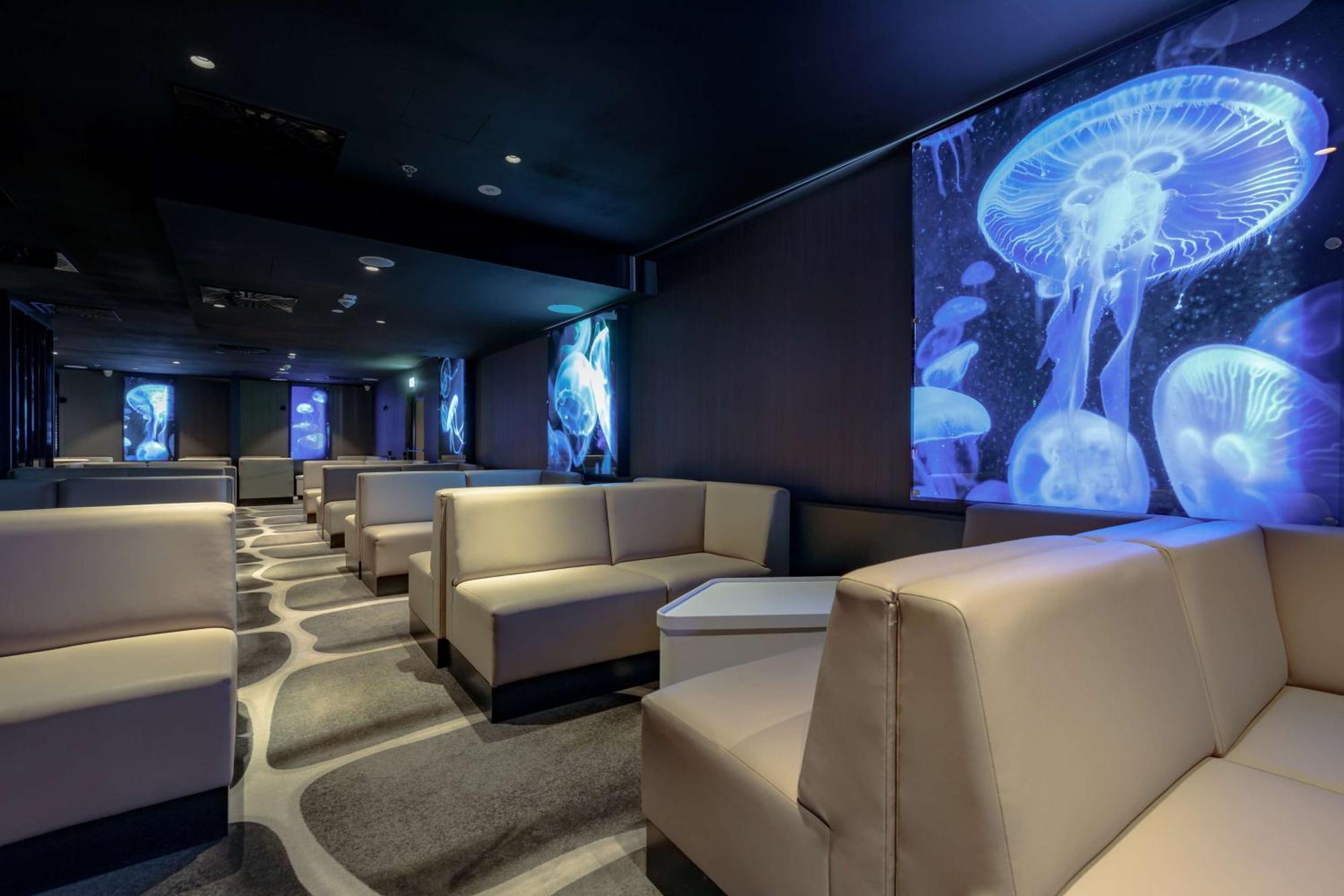 Radisson Blu Resort Swinoujscie Ngoại thất bức ảnh The photo shows a modern lounge space with a sleek design. The area features light-colored, upholstered seating arranged in an open layout. The walls are decorated with large, vibrant images of jellyfish illuminated in blue, creating a calming underw