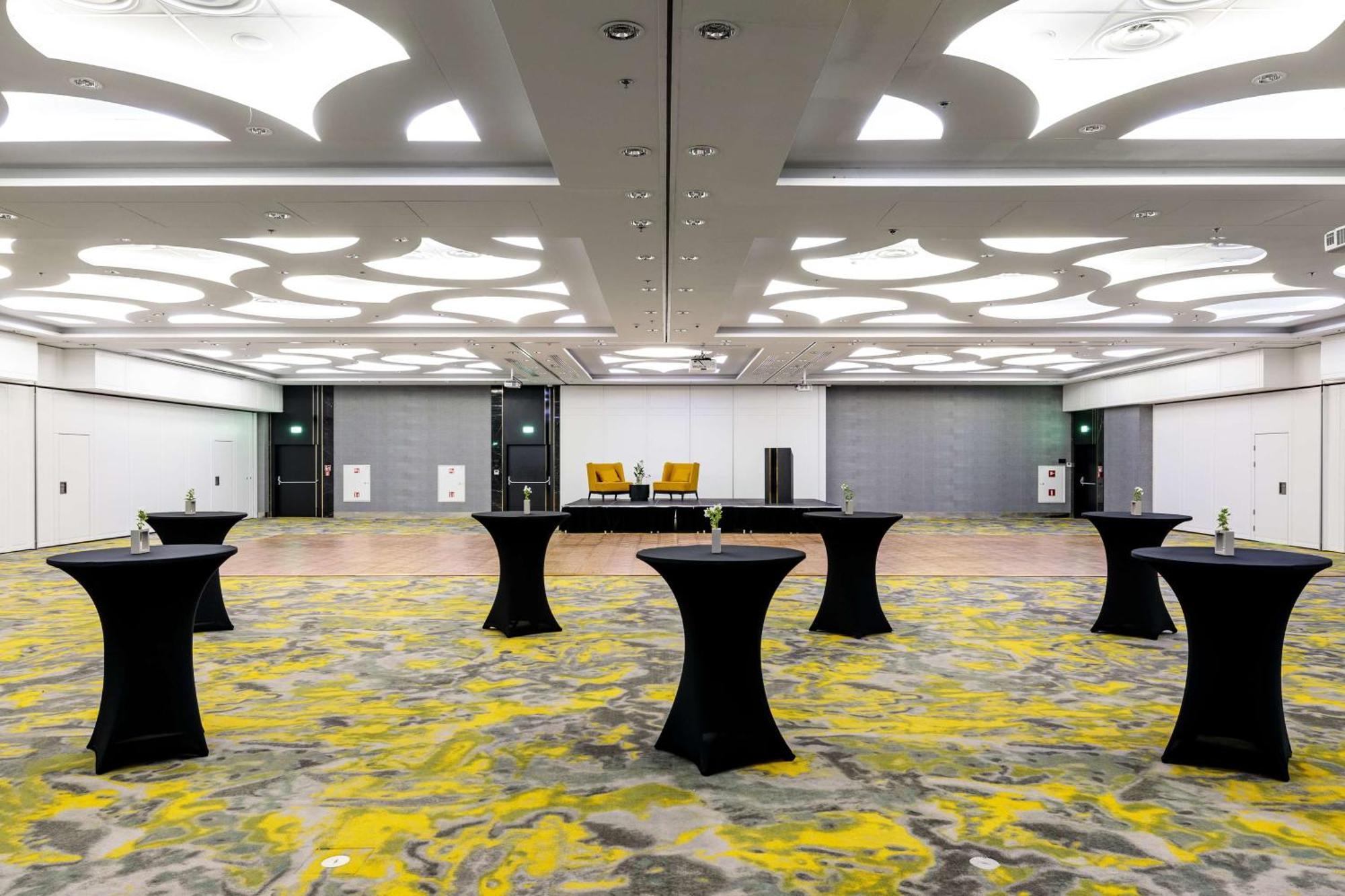 Radisson Blu Resort Swinoujscie Ngoại thất bức ảnh The photo shows a spacious interior room set up for an event. The flooring features a patterned design in gray with yellow accents. There are several black tall tables scattered throughout the area, and in the background, there are two large yellow a