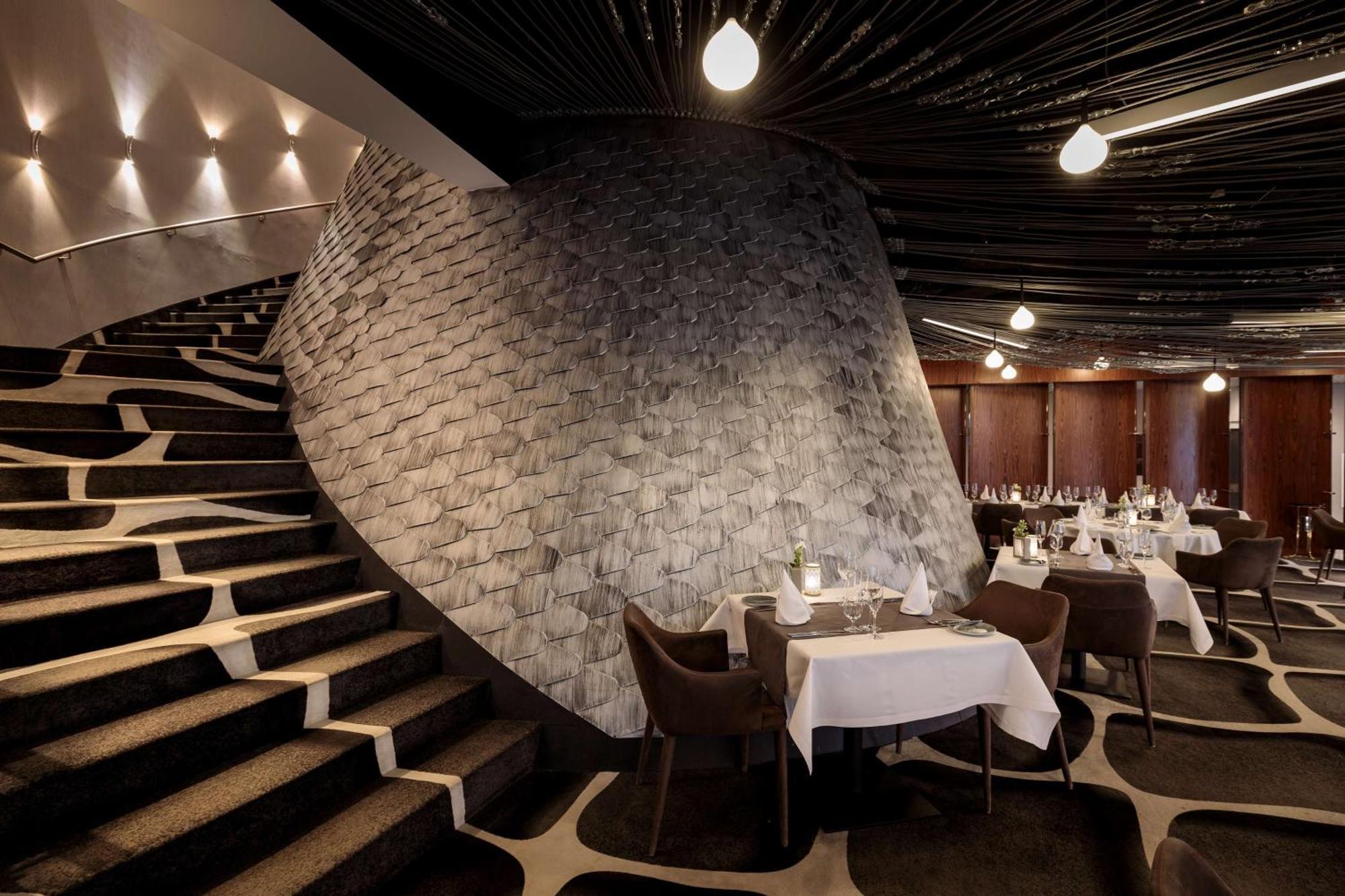 Radisson Blu Resort Swinoujscie Ngoại thất bức ảnh The photo shows the interior of a stylish restaurant. In the foreground, there are elegantly set tables with white tablecloths and dark chairs. A unique, curving stone wall adds a distinct architectural feature, and a grand staircase with a textured 