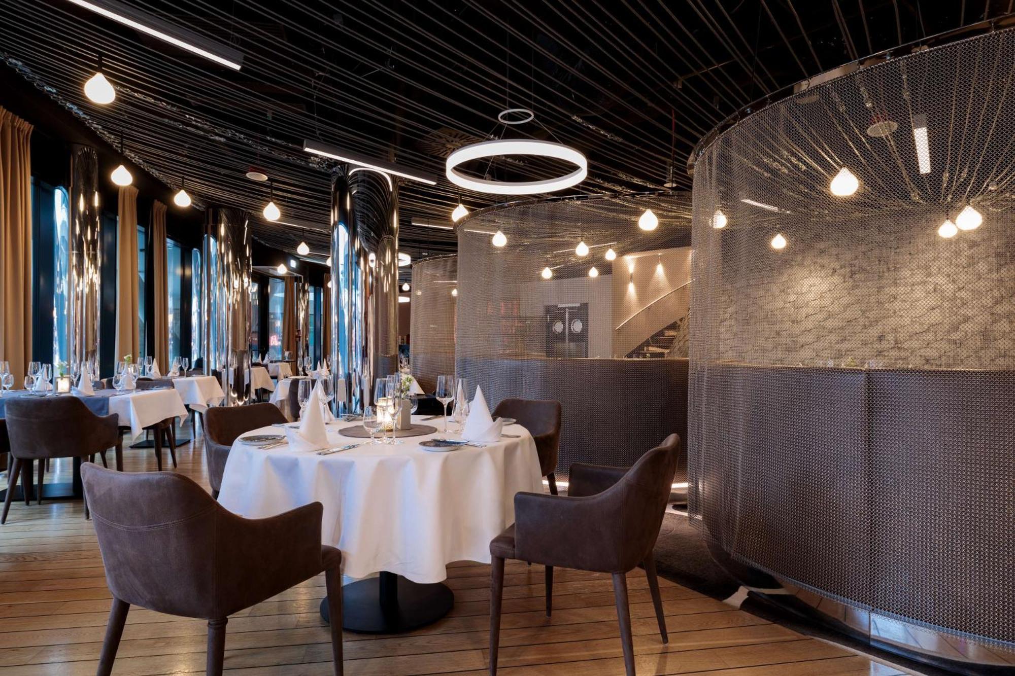 Radisson Blu Resort Swinoujscie Ngoại thất bức ảnh The image shows a modern restaurant interior that features a stylish design. There are several round tables with white tablecloths. The seating consists of brown, upholstered chairs. The space is illuminated by various hanging lights, including a dis
